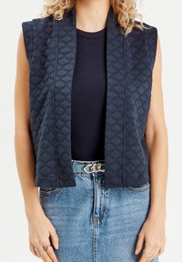 Gilet sans manches en tissu matelassé bleu marine avec un motif texturé, design ouvert à l'avant, associé à un haut sombre et une ceinture bleu clair sur une jupe en jean.