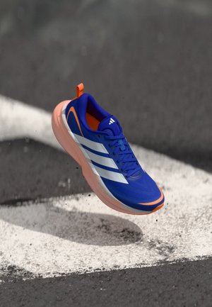 Chaussure de sport bleue avec des rayures blanches, des accents orange, une tige texturée et une semelle légère, flottant au-dessus d'une surface peinte.