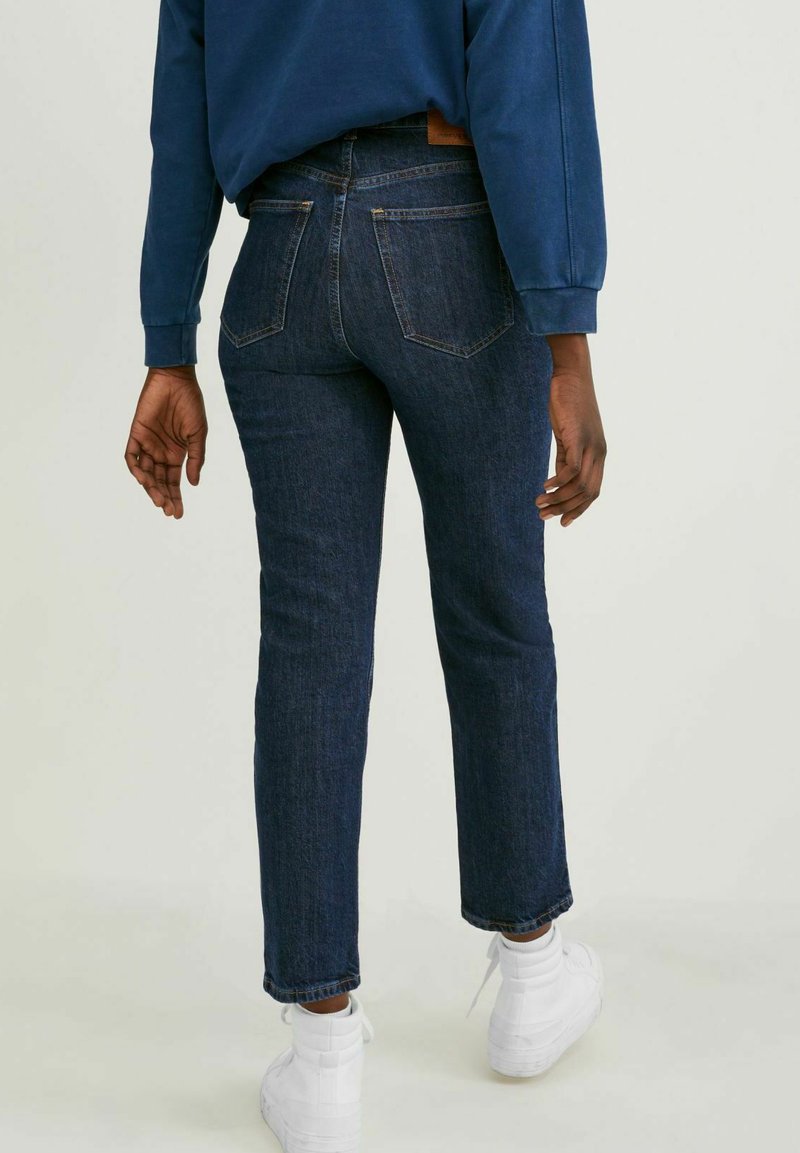 Personne portant un jean bleu foncé, des baskets blanches montantes et un sweat-shirt bleu, se tenant de dos face à l'appareil photo sur un fond uni.