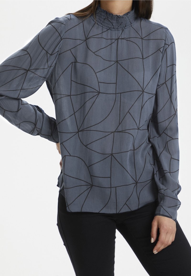 Femme portant un chemisier gris à manches longues avec des lignes noires abstraites, un col montant froncé et un pantalon noir, debout, main sur la hanche.