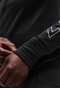 Roke, ki prilagajajo rokav črne športne majice z dolgimi rokavi in belim geometričnim logotipom na podlakti.