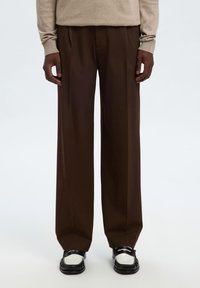 Άτομο που φοράει καστανό παντελόνι με ευρύ πόδι, μπεζ πουλόβερ και διχρωμία μαύρα και άσπρα μοκασίνια, στέκεται μπροστά σε λευκό φόντο.