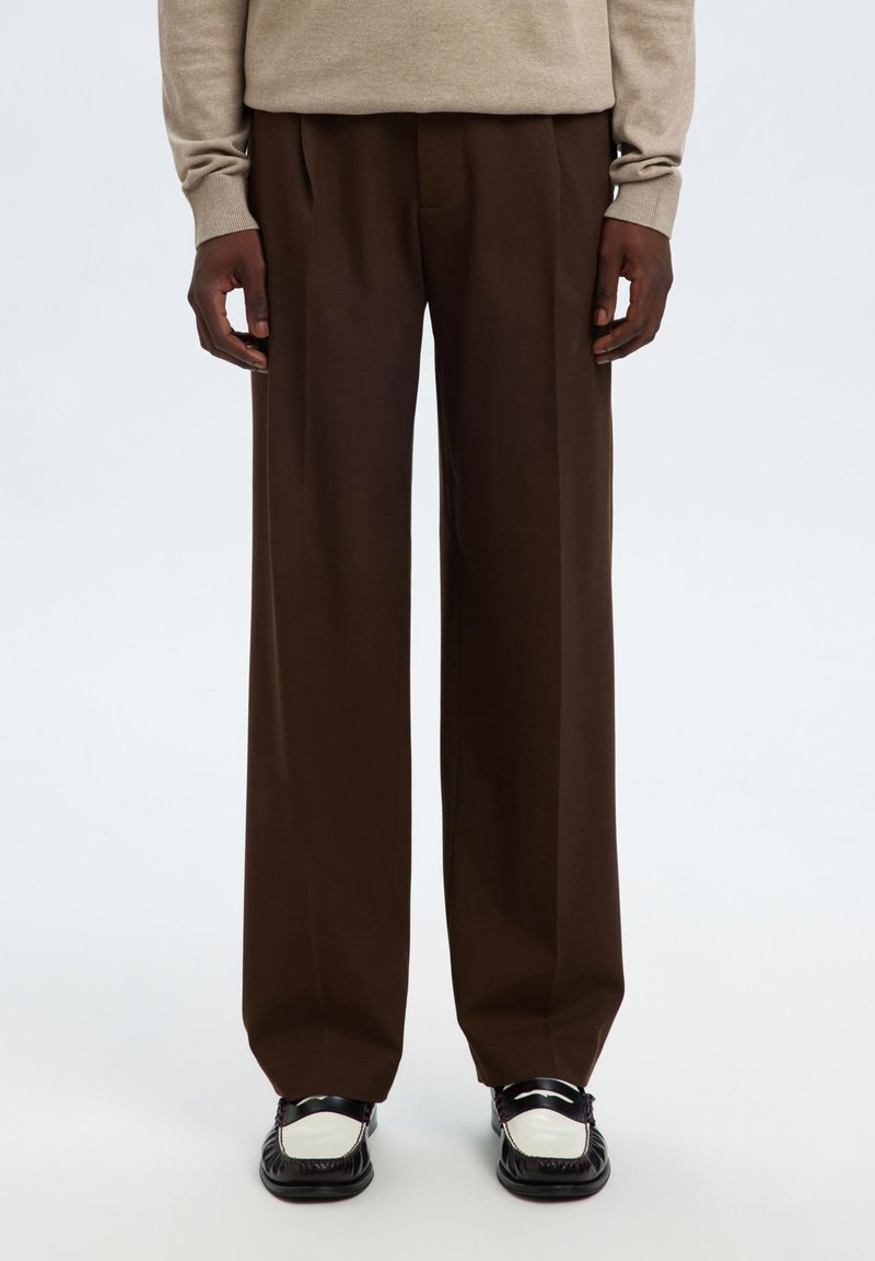 Άτομο που φοράει καστανό παντελόνι με ευρύ πόδι, μπεζ πουλόβερ και διχρωμία μαύρα και άσπρα μοκασίνια, στέκεται μπροστά σε λευκό φόντο.