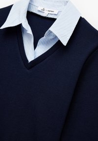 Granatowy sweter z dzianiny z dekoltem w kształcie litery V, nałożony na jasnoniebieską koszulę w białe paski z kołnierzykiem. Metka widoczna przy dekolcie.