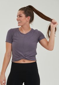 Lila ribbad kort topp med en vriden framdekoration, kombinerad med högmidjade svarta leggings. Modell ler och håller håret i en hästsvans.