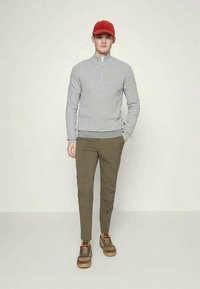 Grijze rits trui van textiel, gecombineerd met olijfgroene slim-fit broek en bruine schoenen met rode en gele accenten. Rode pet.