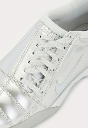 Baskets en cuir métallisé blanc avec lacets blancs, coutures apparentes, col rembourré et discret swoosh Nike sur le côté.