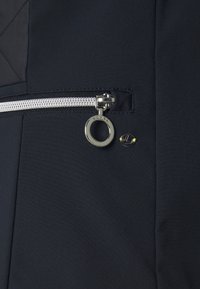 Tkanina kurtki w kolorze granatowym z srebrnym zamkiem błyskawicznym, okrągłym uchwytem zamka i małym logo marki. Posiada teksturowany wzór i eleganckie linie.