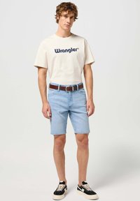Beige t-shirt met korte mouwen van het merk Wrangler, gecombineerd met lichtblauwe spijkerbroekshorts en een bruine leren riem. Zwarte sneakers met witte zolen.