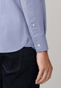 Mano di un uomo appoggiata accanto a jeans scuri e a una manica di camicia blu con bottoni, caratterizzata da un piccolo motivo a quadretti e dettagli con orlo arrotondato.