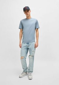 T-shirt azzurro chiaro a maniche corte, jeans in denim leggero strappati, e sneakers bianche. Cappellino grigio con logo rosso "HUGO".