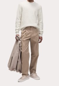 Beige byxor med veck, vit strukturerad tröja och ljusa sneakers, håller en beige jacka. Enkel, modern design.