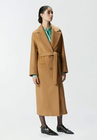 Dugi smeđi kaput s remenom, velikim reverima, dva prednja džepa i smešnim dugmadi. Nosi se preko zelene košulje s uzorkom i uparen je s crnim cipelama.