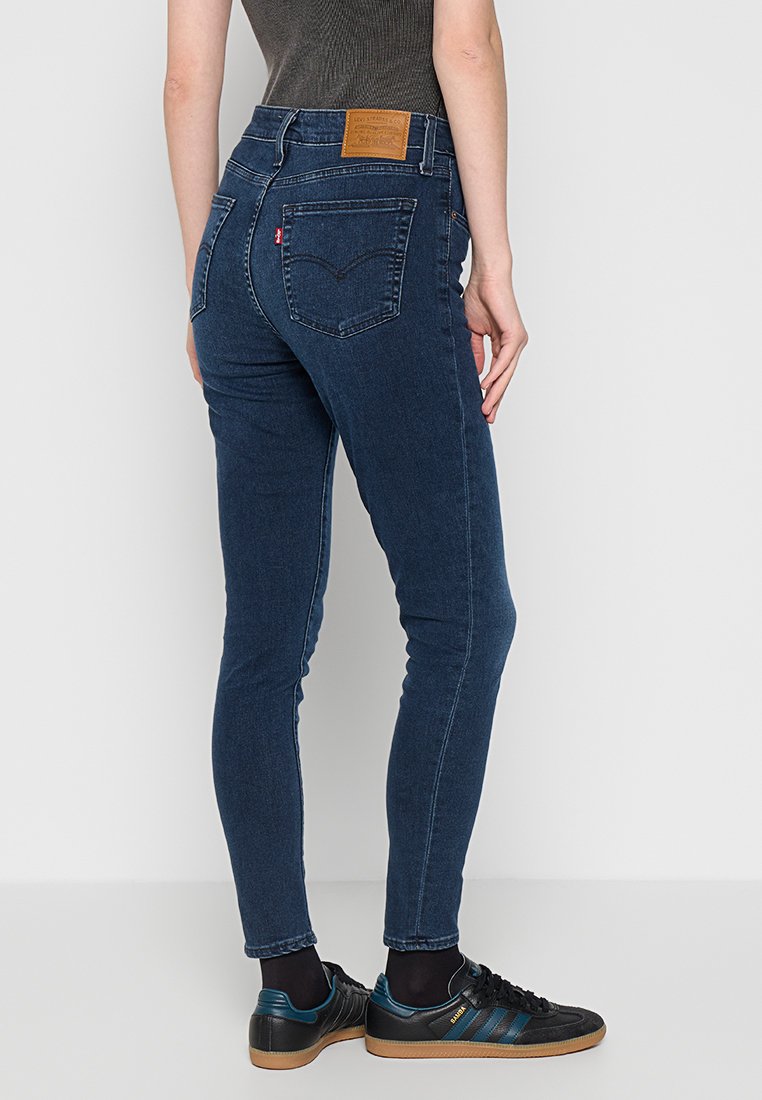 Eine Person trägt dunkelblaue Levi's Skinny Jeans, schwarze Socken und schwarze Adidas Sneakers mit blauen Streifen und steht vor einem schlichten weißen Hintergrund.