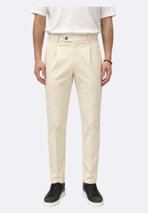 Uomo che indossa pantaloni beige plissettati, t-shirt bianca, braccialetto nero e sneakers nere con suola bianca, in piedi contro uno sfondo semplice.