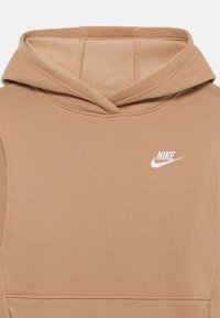 Hanorac cu glugă din material bej, având o textură subtilă. Un logo alb Nike este amplasat pe piept. Detaliile cusăturii sunt vizibile pe glugă.