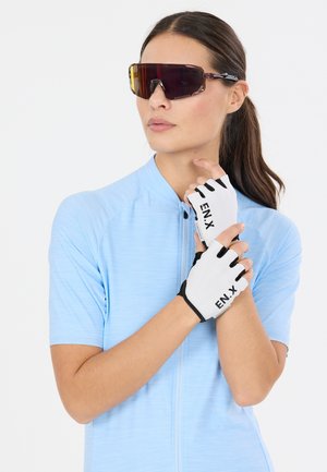 Vrouw in een lichtblauw wielershirt met witte vingervrije handschoenen en donkere sports zonnebril met omhulconcept, die een handschoen aan haar linkerhand verstelt.