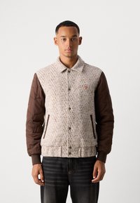 Giovane uomo che indossa una giacca di lana beige con maniche trapuntate marroni e jeans scuri, rivolto in avanti contro uno sfondo chiaro e semplice.
