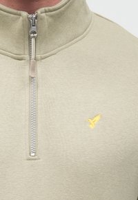 Heleroheline lukuga dressipluus kõrge kraega, millel on hõbedane lukk ja vasakul rinnal väike kollane tikitud linnu logo.