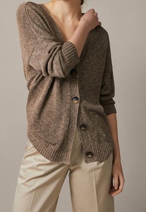Personne portant un cardigan marron boutonné avec les manches retroussées et un pantalon beige taille haute, debout devant un fond uni.