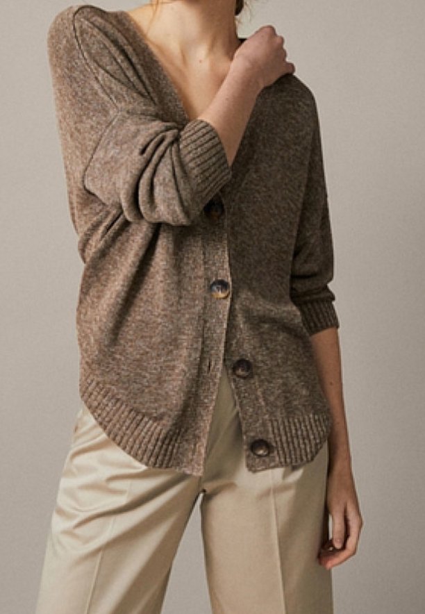 Personne portant un cardigan marron boutonné avec les manches retroussées et un pantalon beige taille haute, debout devant un fond uni.