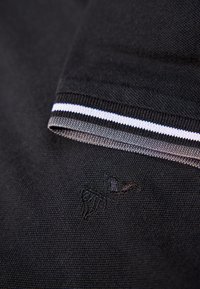 Nära vy av ihopfällt svart tyg med vit och svart randig kant och subtilt broderad logotyp synlig.