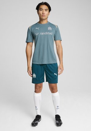 Maillot vert-bleu à manches courtes avec un motif texturé et un logo. Shorts vert-bleu foncé, chaussettes blanches avec des accents bleus, et chaussures de soccer noires.