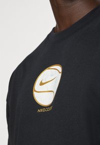 Must musta T-särk, millel on suur valge ja kuldne Nike logo lillemustriga ning allpool logo kuldne tekst "NIKE COURT".