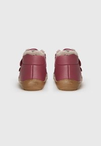 Par de zapatos de cuero rojo para niños pequeños con forro de felpa beige, costuras visibles y suelas de goma marrón, vistos desde la parte trasera sobre un fondo liso.