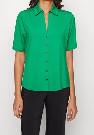 Vrouw met een groen overhemd met korte mouwen en een kraag, gecombineerd met een zwarte broek, staande tegen een eenvoudige achtergrond.