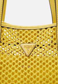 Sac en bandoulière tissé jaune avec un design en maille texturée, arborant un logo Guess en métal doré en forme de triangle et des poignées en cuir durable.