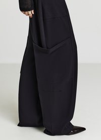 Schwarze, weit geschnittene Hosen aus glattem Stoff, mit großen Seitentaschen und lockerem Sitz. Am unteren Rand sind spitze schwarze Schuhe zu sehen.