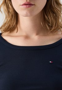 Dunkelblaues geripptes Oberteil mit weitem Ausschnitt und kurzen Ärmeln. Verfügt über ein kleines gesticktes Logo nahe der linken Schulter. Weiche Textur.