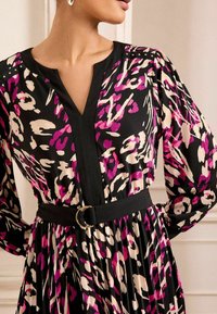 Duga haljina dugih rukava u crnoj boji s apstraktnim uzorkom u ružičastoj i krem boji, s V-izrezom, crnim pojasom sa zlatnim detaljima i plisiranom suknjom.