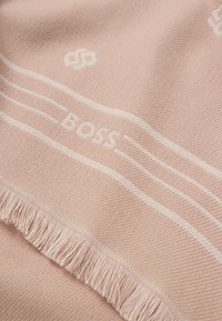 Jasnoróżowy pleciony koc z białymi paskami i napisem "BOSS". Krawędzie są frędzlowane, co dodaje tekstury i szczegółowości do projektu.