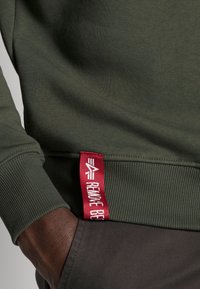 Groene sweatshirt met ribgebreide boorden en zoom, met een rood label waarop "REMOVE BE" staat. Soepele textuur, nauwsluitend ontwerp en ronde halslijn.