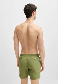 Green swim shorts with elastic waistband, single back pocket with zipper, and a smooth fabric texture. Model is shown from the back.