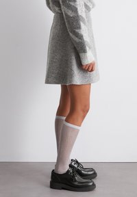 Persoon in een middellange grijze rok, een gemêleerd grijs sweater, lichtgrijze kniekousen en zwarte veterschoenen die op een gladde vloer staat.