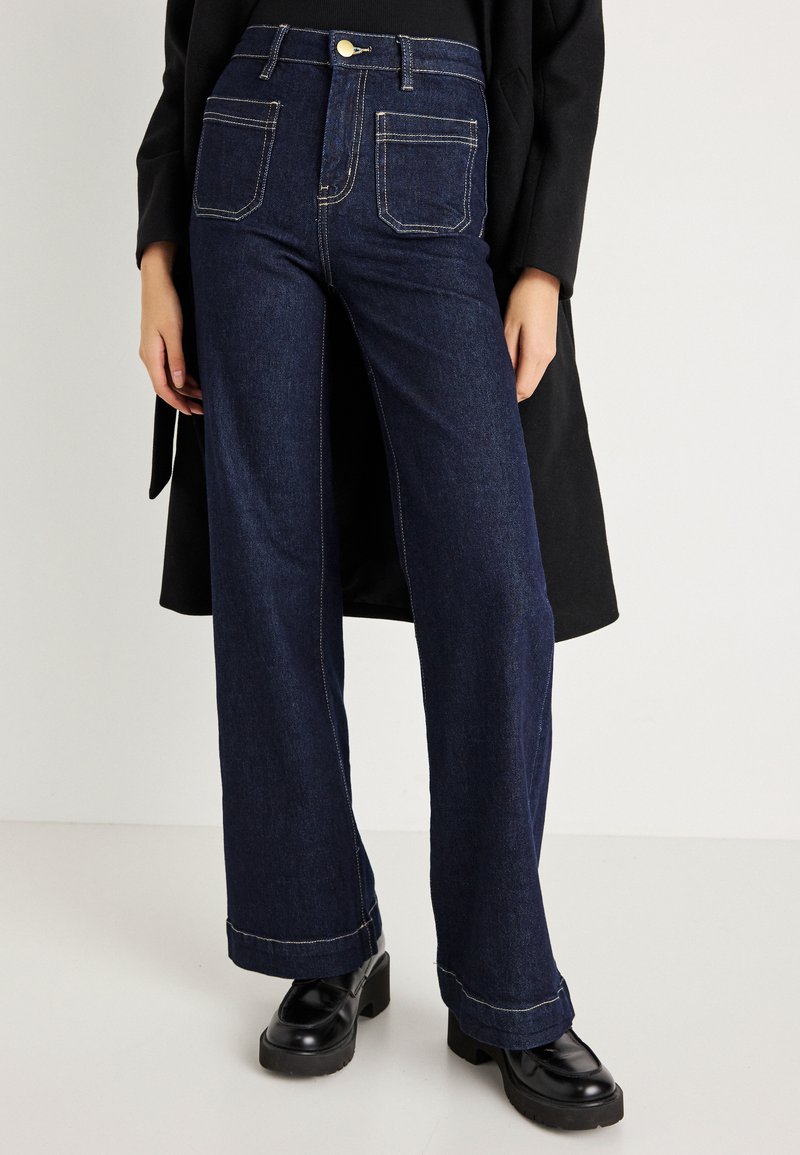 Tumši zili plata kāju džinsi, izgatavoti no denima, ar augstu jostasvietu, priekšējo pogu aizdari un baltu šuvi detaļām uz kabatām.