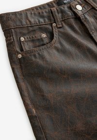 Jeans de denim marrón oscuro con un acabado texturizado, que presentan un bolsillo frontal, costuras en contraste y detalles de botones metálicos.