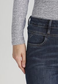 La mano izquierda descansando junto a la cadera, vistiendo jeans de mezclilla azul oscuro y una camiseta de manga larga gris claro, contra un fondo simple.