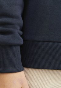 Sötétkék kapucnis pulóver mandzsettája bordázott textúrájú, sima felülettel. Látható kéz egy világos színű kordbársony anyagnak támaszkodik.