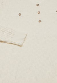 Cardigan crem cu un model de tricotat texturat, având cinci butoane decorative și două buzunare pe piept cu închidere cu buton. Mâneci texturate.
