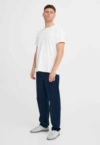 Witte katoenen t-shirt gecombineerd met marineblauwe broek, met een ontspannen pasvorm. De persoon draagt grijze sneakers met witte strepen en staat tegen een eentonige achtergrond.