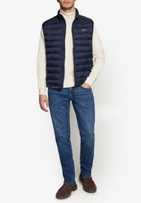 Mørkeblå quiltet vest med høj krave, lynlås foran, båret over en lys turtleneck. Kombineret med blå jeans og brune sko.