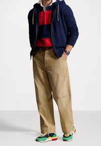 Marinblå zip-hoodie över en röd och marinrandig polo, tan vida byxor med synliga sömmar, och flerfärgade sneakers med en kraftig sula.