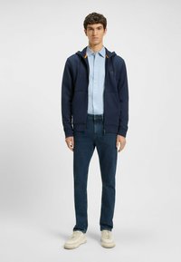 Marineblå zip-up hettegenser med oransje interiørdetaljer, brukt over en lyseblå button-up skjorte, kombinert med mørke jeans og kremfargede joggesko.