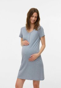 Gravid kvinna i en kortärmad blå klänning med vita mönster, som försiktigt håller om sin mage och ler medan hon ser ner.