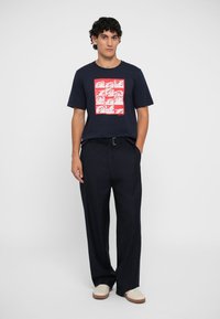 Camiseta de algodón azul marino con un diseño gráfico rojo, combinada con pantalones oscuros de rayas finas y zapatos de color claro, de pie sobre un fondo blanco.