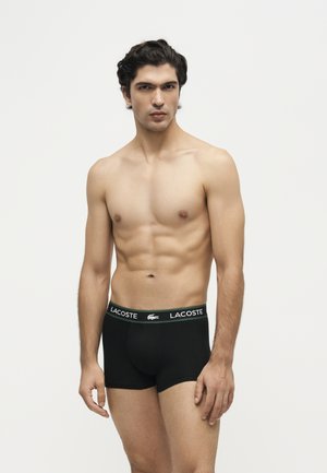 Jeune homme aux cheveux foncés portant un boxer noir Lacoste, debout sur un fond blanc uni.